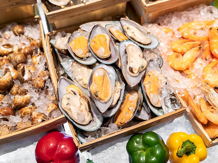 A tray of fresh oysters on ice, with shells open and orange roe visible, surrounded by other shellfish and colorful bell peppers.
