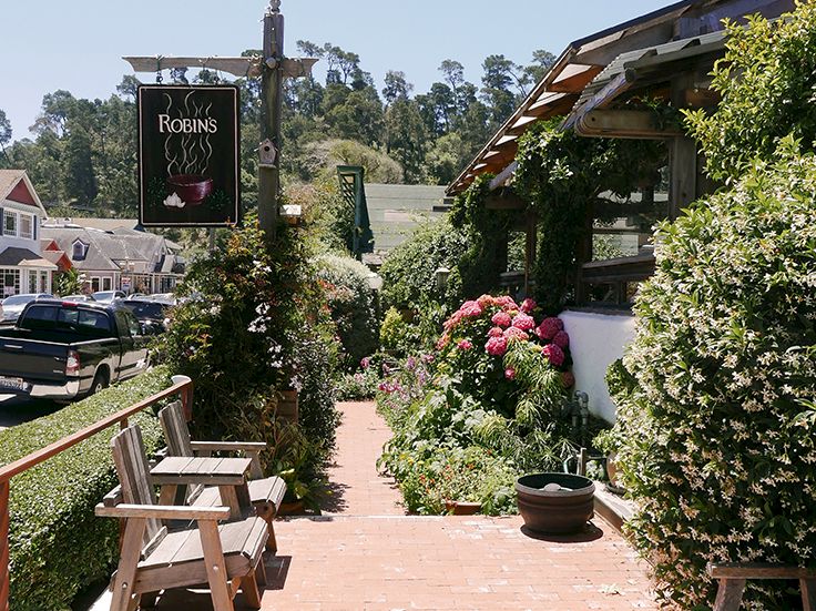 A quaint outdoor porch scene with flower-filled path, wooden seating, a hanging sign for &ldquo;Robins,&rdquo; and a sunlit village vibe.