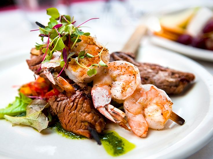 Sliced steak with shrimp tails on a bed of greens, drizzled with green sauce and a garnish of microgreens, plated artfully.