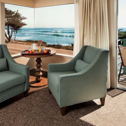 A cozy sea-view lounge with two teal armchairs, a small round table, and a balcony overlooking the ocean. 140 chars, ending with a period.