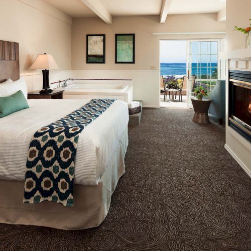 A cozy hotel room with a king bed, patterned throw, neutral tones, fireplace, and a door to a balcony overlooking water.