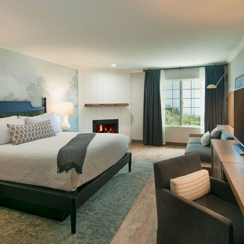 A spacious hotel room with a large king bed, sitting area, fireplace, desk, and a window with blue curtains, all in neutral tones.
