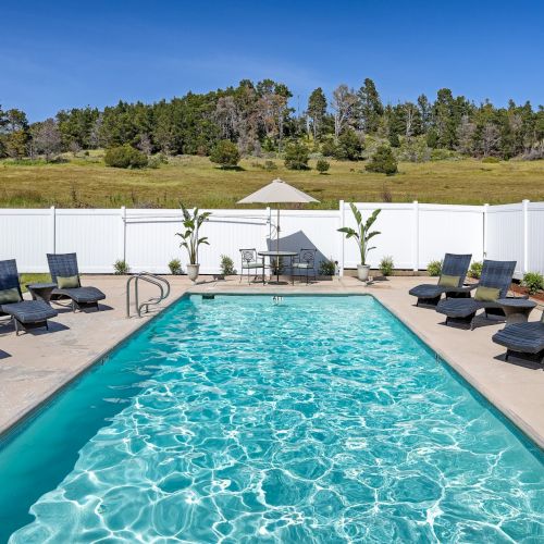 A bright rectangular backyard pool with clear blue water, surrounded by black lounge chairs, a white fence, and a sunny lawn beyond.