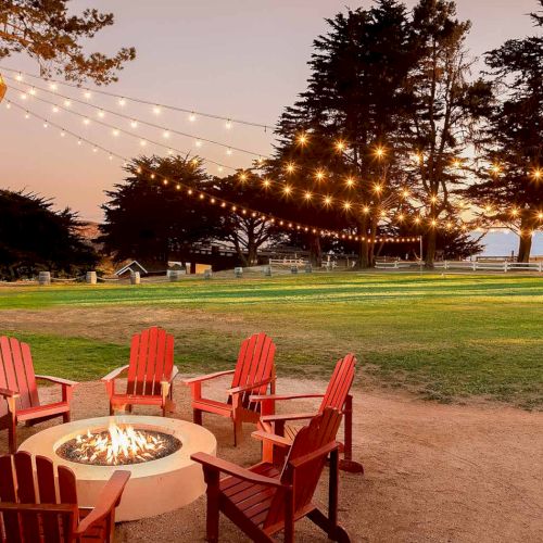 A cozy outdoor gathering by a fire pit with red chairs, string lights, trees, and a grassy park at dusk, perfect for socializing and warmth.
