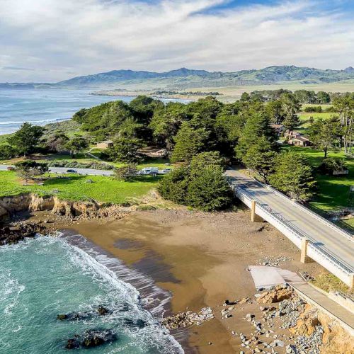 A coastal cliffside with a road crossing a small bridge over a sandy cove, green fields, and pine trees, meeting blue ocean under a cloudy sky.