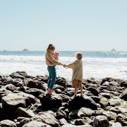 Two people holding hands on a rocky shore, waves crash behind them under a clear sky. Safe moment of connection by the sea.