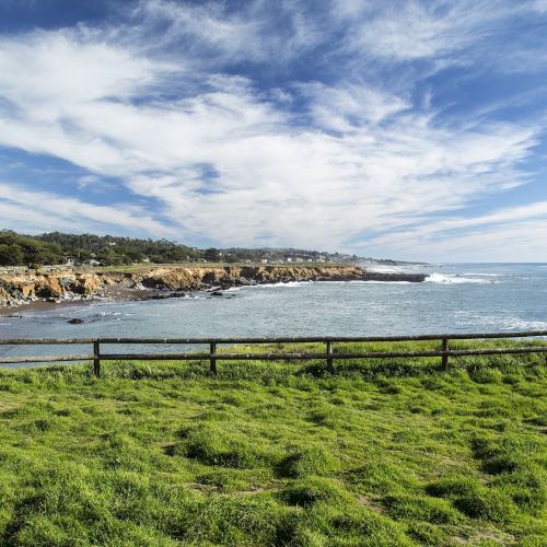 A rocky coastline with green grass, a wooden fence, and calm blue sea under a partly cloudy sky.