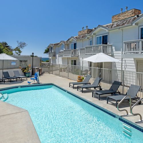 A bright outdoor pool area surrounded by lounge chairs and umbrellas, with a two-story beige apartment building in the background and a clear blue sky.