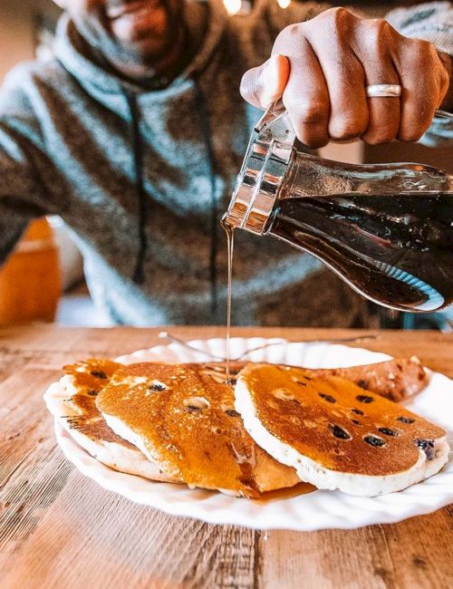 Someone pours syrup over a stack of pancakes on a wooden table, drizzle dripping onto the plate.