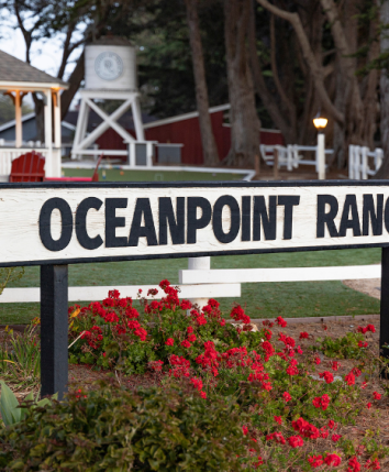 Sign reads &ldquo;Oceanpoint Ranch&rdquo; at front gate, with white fences, red chairs, gazebo, and floral garden surrounding the entrance.
