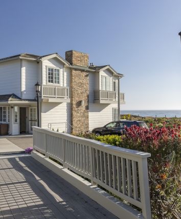 Two-story beachfront house with stone chimney, white siding, a gated path, a driveway, and a lamppost overlooking the ocean under a sunny sky.