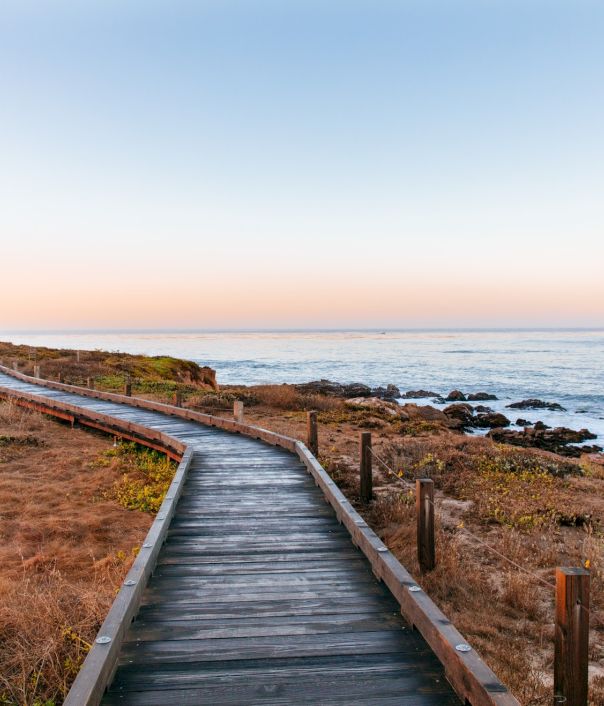 A wooden boardwalk winds toward the sea along a rocky coastal path at sunset, with dry grass, posts, and calm waves.