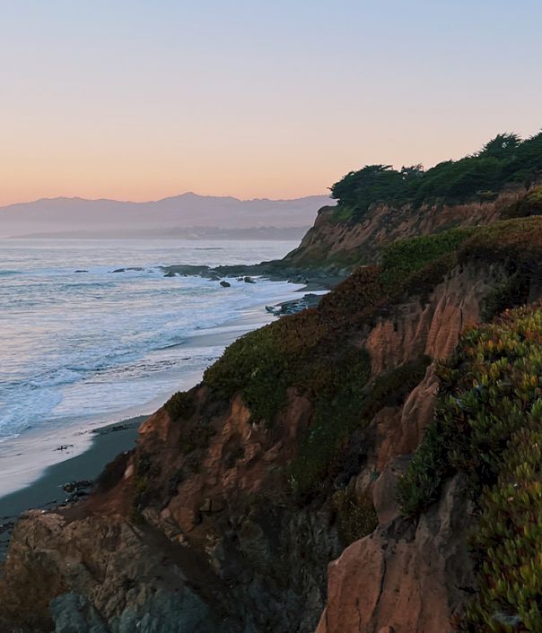 Coastal cliffside at sunset, waves crash on a rocky shore, evergreen brush covers the hillside, warm sky fades over distant hills.