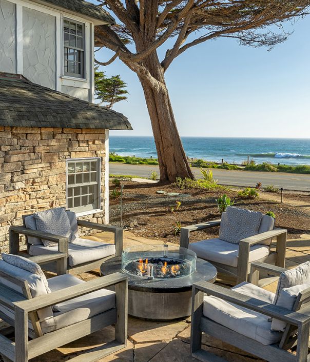 Cozy outdoor patio with gray cushioned chairs around a circular fire pit, stone house wall, trees, and a beachfront view by the ocean.