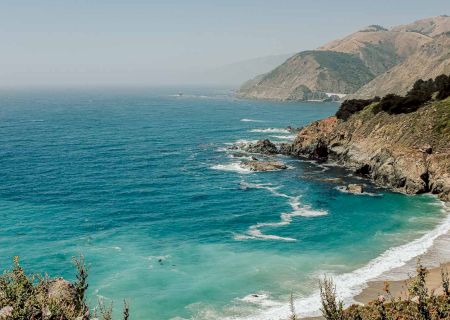 A scenic coastline with turquoise waves crashing against a rocky shore, hills in the distance, and a clear horizon over the calm sea.